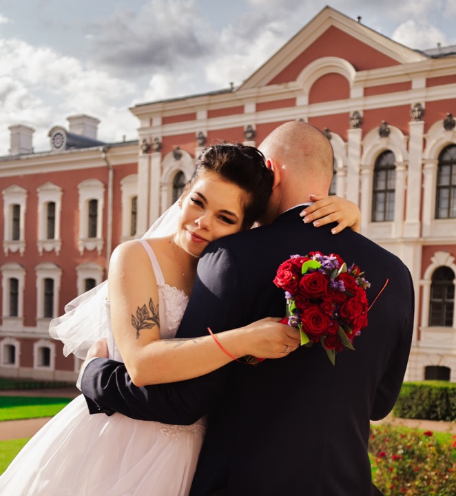 A bride with a red bouquet looking towards the camera over the groom’s shoulder